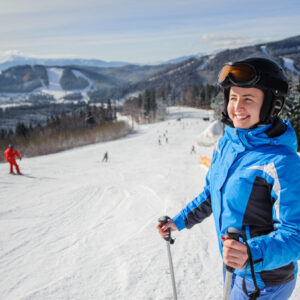 Close-up portrait of young happy woman skier looking at beautiful mountain landscape at the ski resort on a sunny day. Girl is wearing blue jacket helmet and goggles. Wide angle. Bukovel, Ukraine